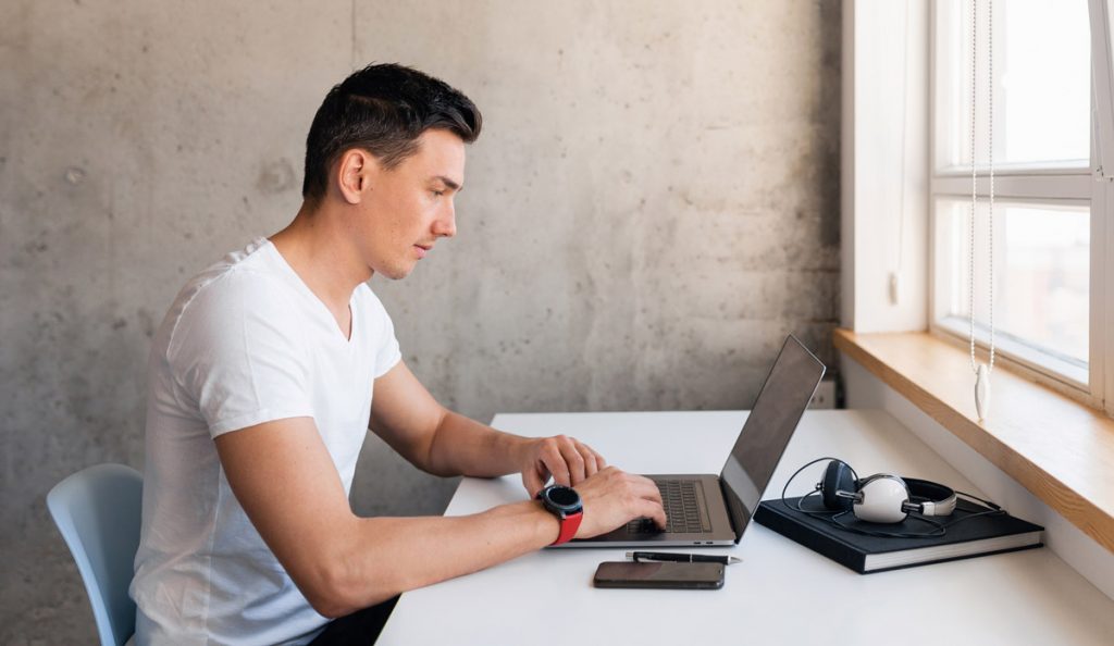 young handsome smiling man in casual outfit sitting at table working on laptop staying at home alone, typing, freelancer at work online.