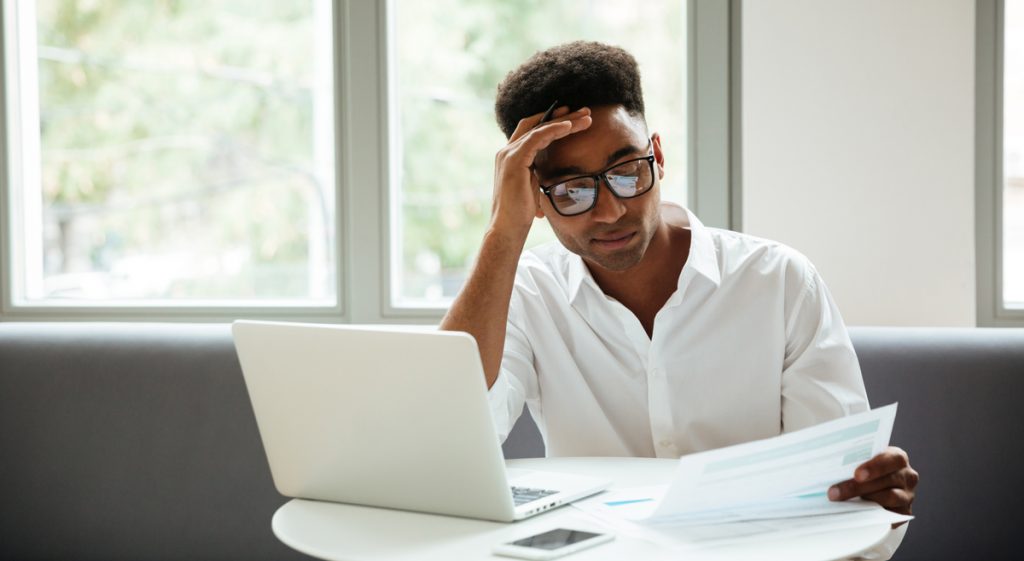 Picture of concentrated serious young african man sitting coworking by laptop computer near documents. Looking aside.