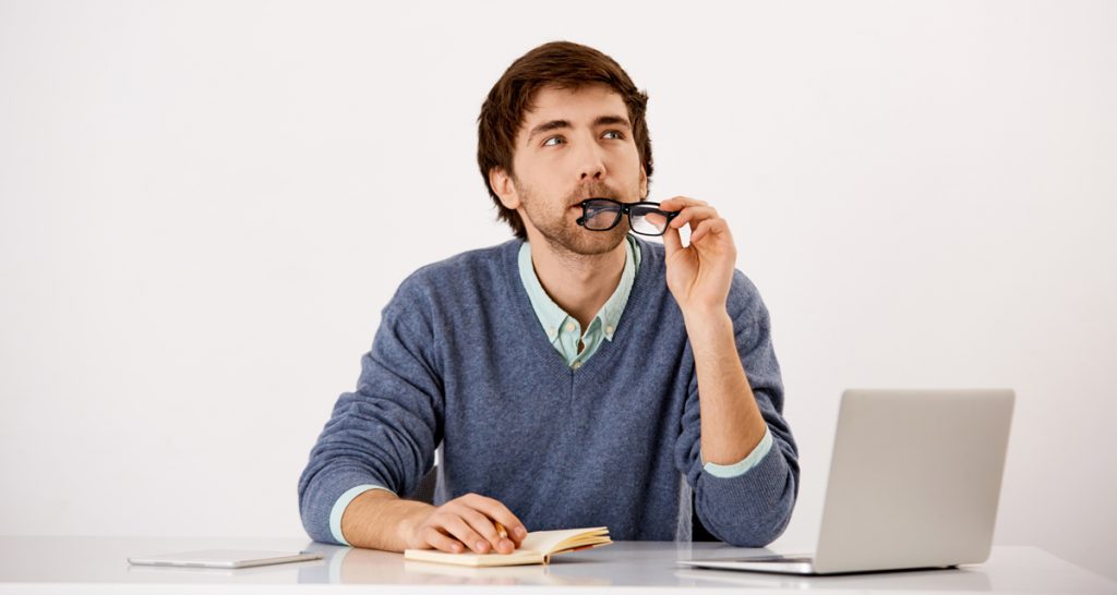 Handsome wondering concentrated young businessman sitting at the office desk writing down something to notebook with a pencil.