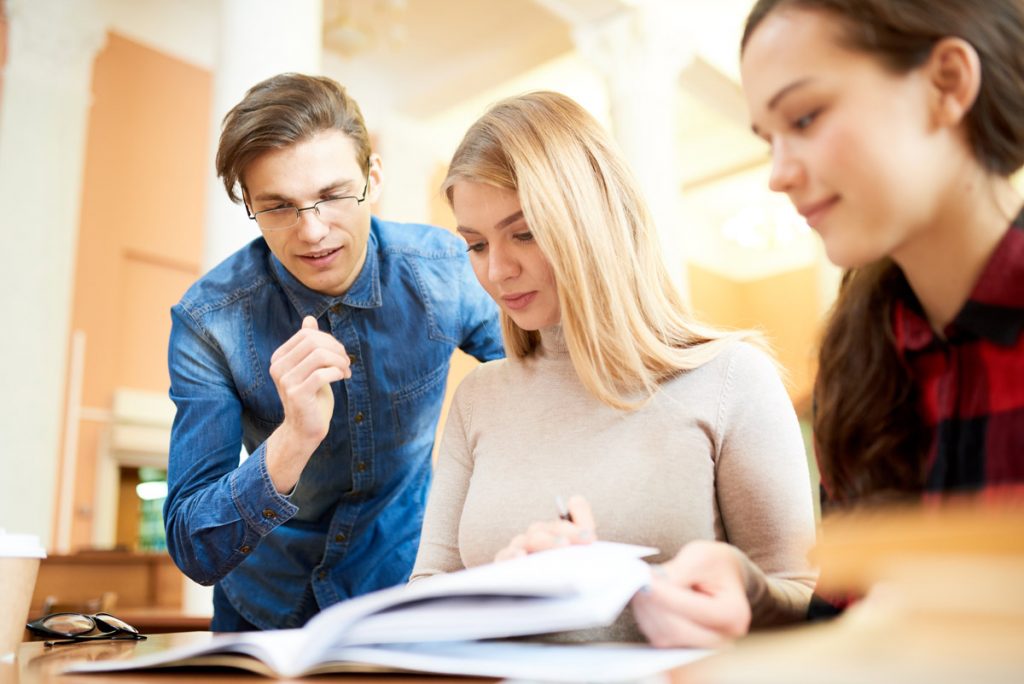 Positive confident handsome young male student explaining difficult topic to concentrated girls in modern library while they studying material together.