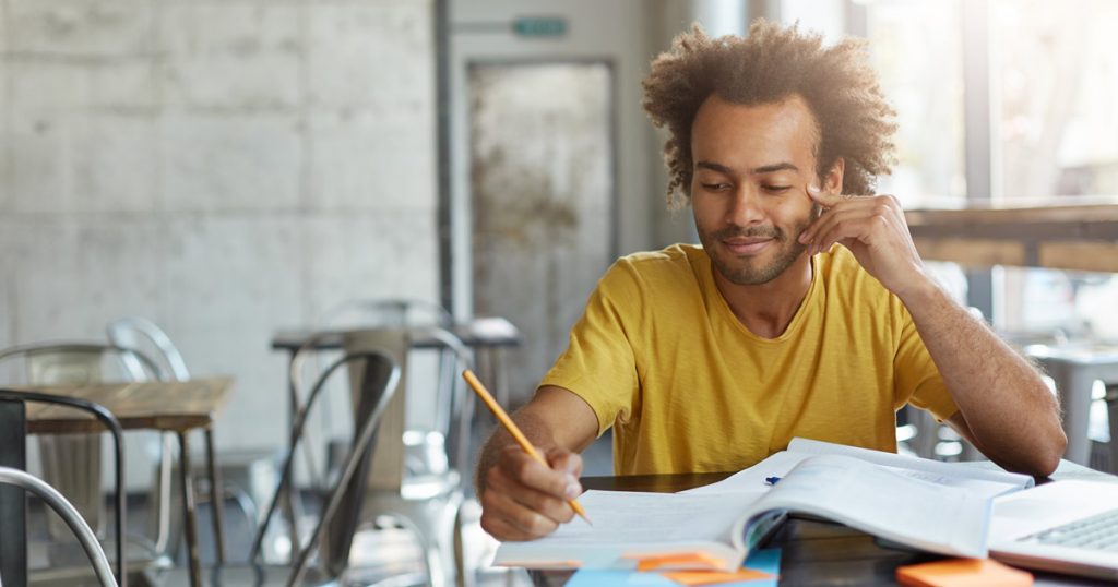 Creative young author with curly hair and dark skin dressed casually sitting at cafeteria preparing for writing new article in his newspaper.