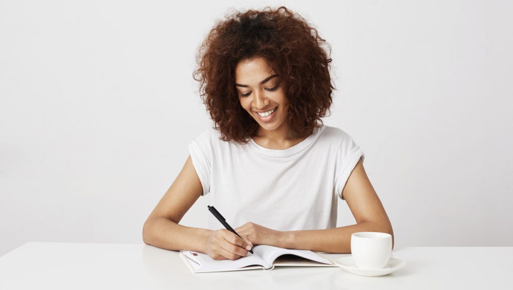 Cheerful african girl smiling writing in notebook at workplace over white background.