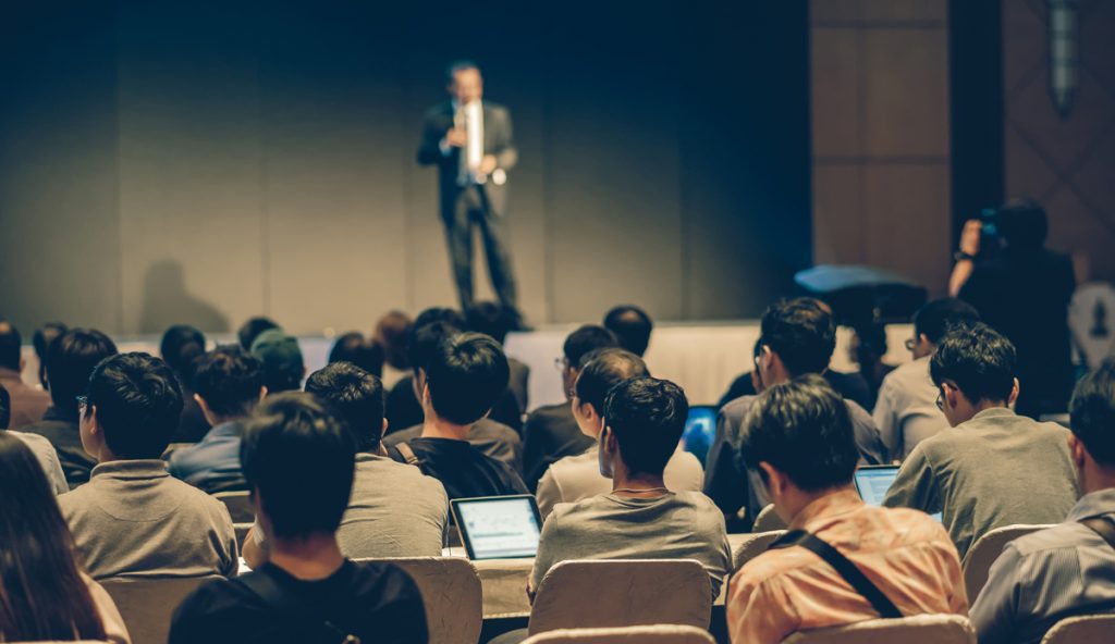 Rear side of Audiences sitting and listening the speackers on the stage in low light conference hall, event and seminar concept.