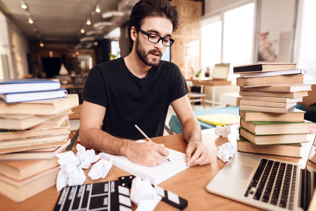 Freelancer bearded man in t-shirt taking notes sitting at desk surrounded by books.