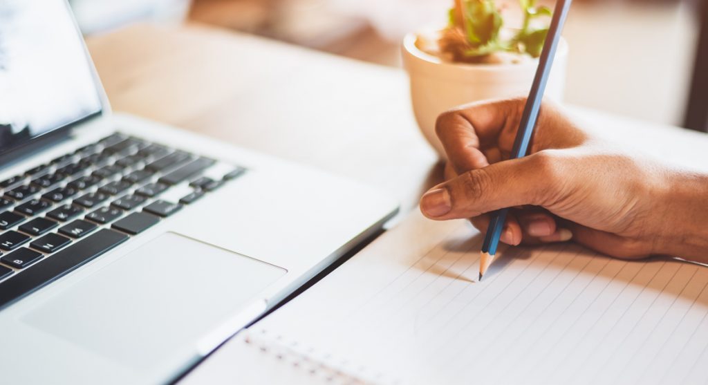 Close up of working woman hand using laptop computer and writing letter on notebook paper in office desk.