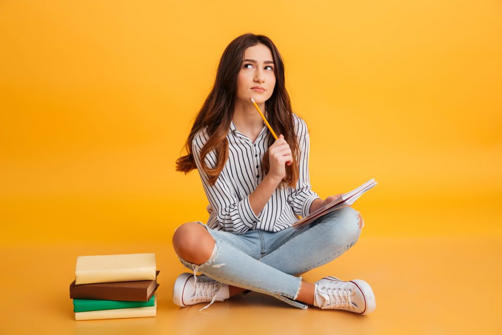 Portrait of a pensive young girl making notes while sitting with books isolated over yellow background.